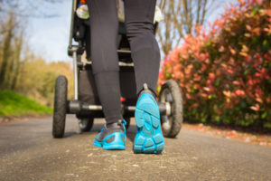 Young mother exercises in a park. Wears blue-gray sport shoes, pushes a pram