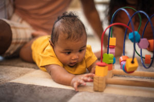 Infant plays with toy on the ground, parents in background.